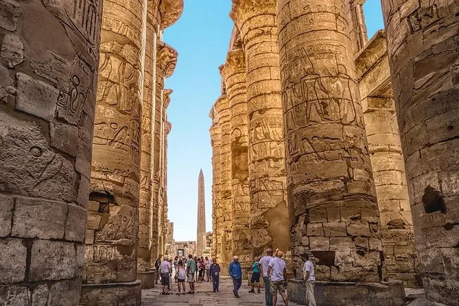 Besucher, die zwischen den mächtigen gemeißelten Säulen der Hypostylhalle von Karnak gehen, ein Obelisk in der Ferne